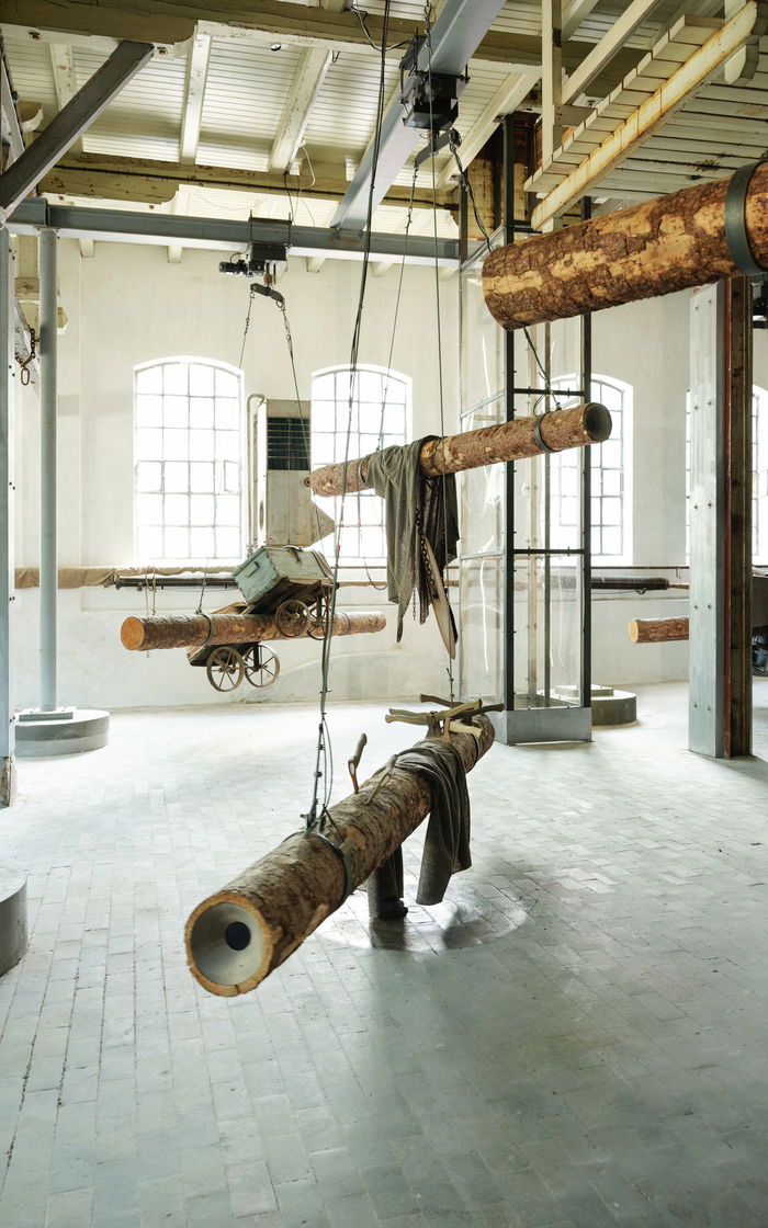 Kunstinstallasjon. Tømmerstokker henger fra taket i en gammel fabrikkhall.